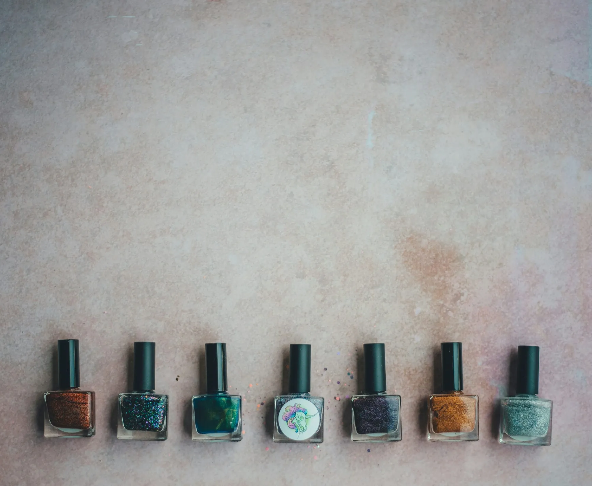 Seven bottles of shimmer nail polish lined up on a pale concrete surface, in copper, teal, violet, gold and silver, with loose glitter scattered between them