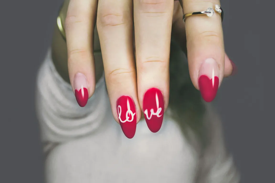 Long almond nails in raspberry red with a deeper red tip and the word love hand-painted in white across two fingers