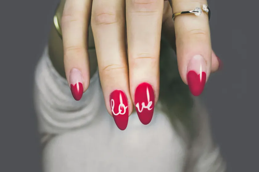 Long almond nails in raspberry red with a deeper red tip and the word love hand-painted in white across two fingers