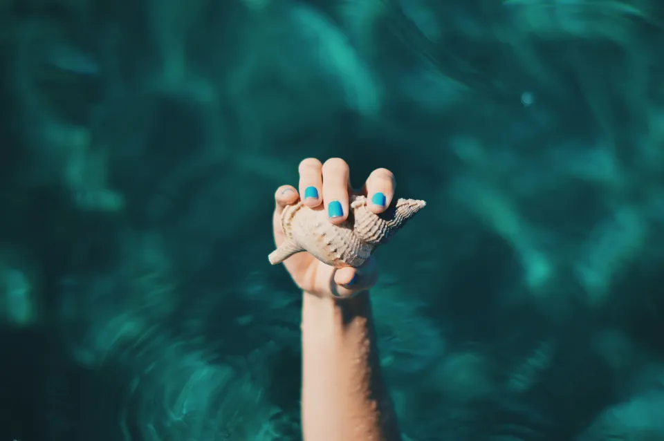 A hand with bright blue nails holding a spiral seashell above clear turquoise water