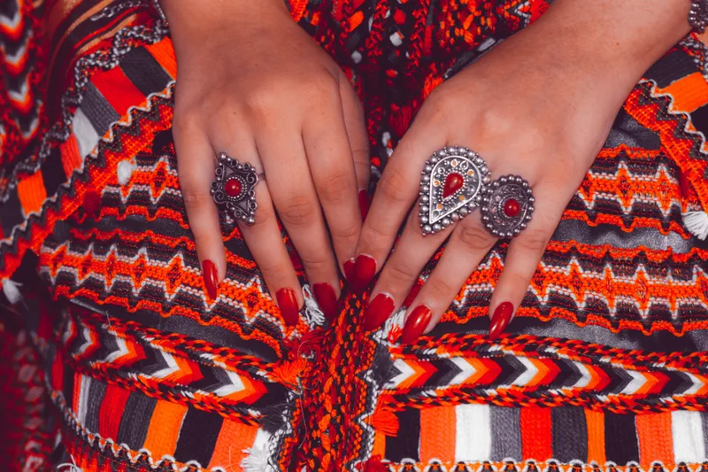 Two hands resting on a red, orange and black woven textile, long nails in glossy brick red, wearing ornate silver rings set with red stones