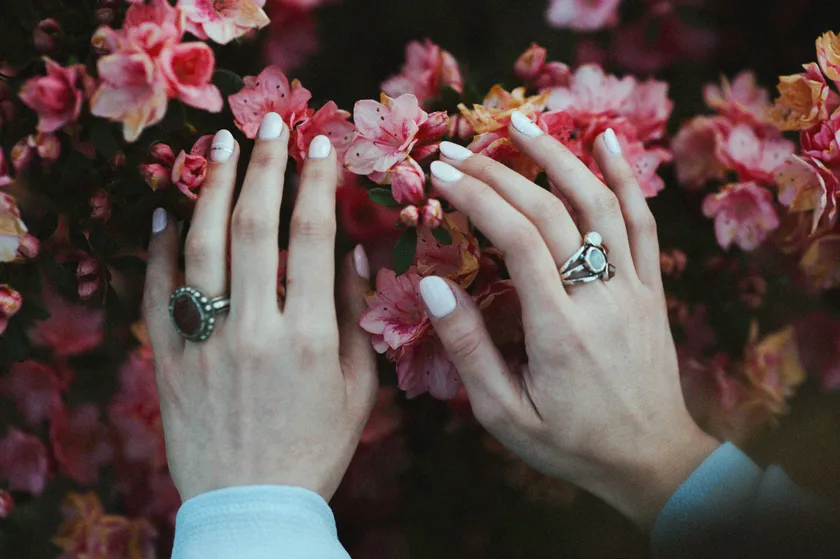 Two hands with short white almond nails resting against a wall of pink azalea blossom