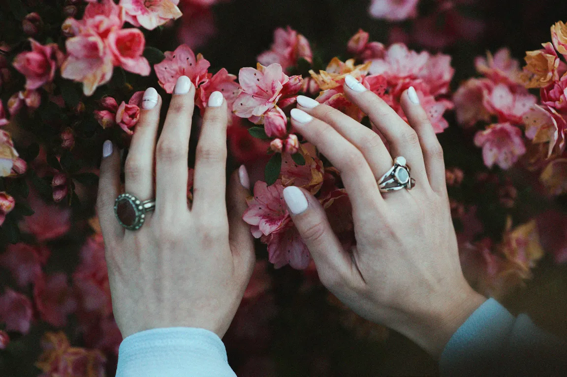 Two hands with short white almond nails resting against a wall of pink azalea blossom