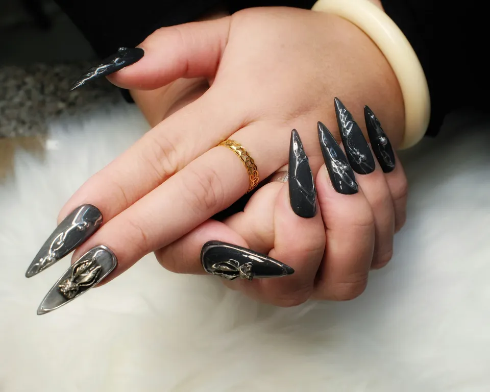 Two hands with long black stiletto nails in glossy silver-marbled chrome, with a gold ring and a cream bangle