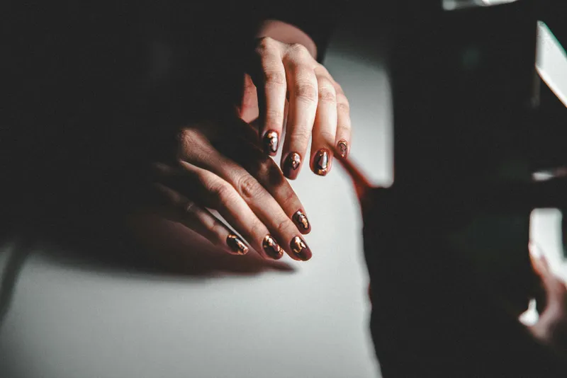 Two hands in low light with short oval nails in deep burgundy, each finished with a leaf of copper foil
