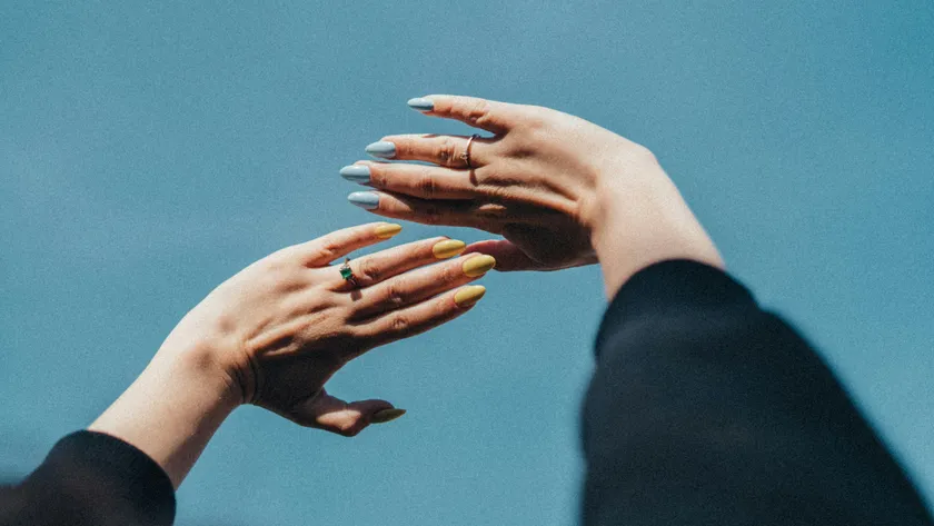 Two hands raised against a blue sky, one set of almond nails in pale blue and the other in lemon yellow