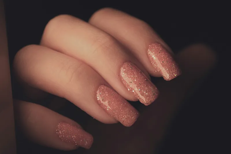 A hand with long square nails in rose pink packed with fine silver glitter, against a dark background