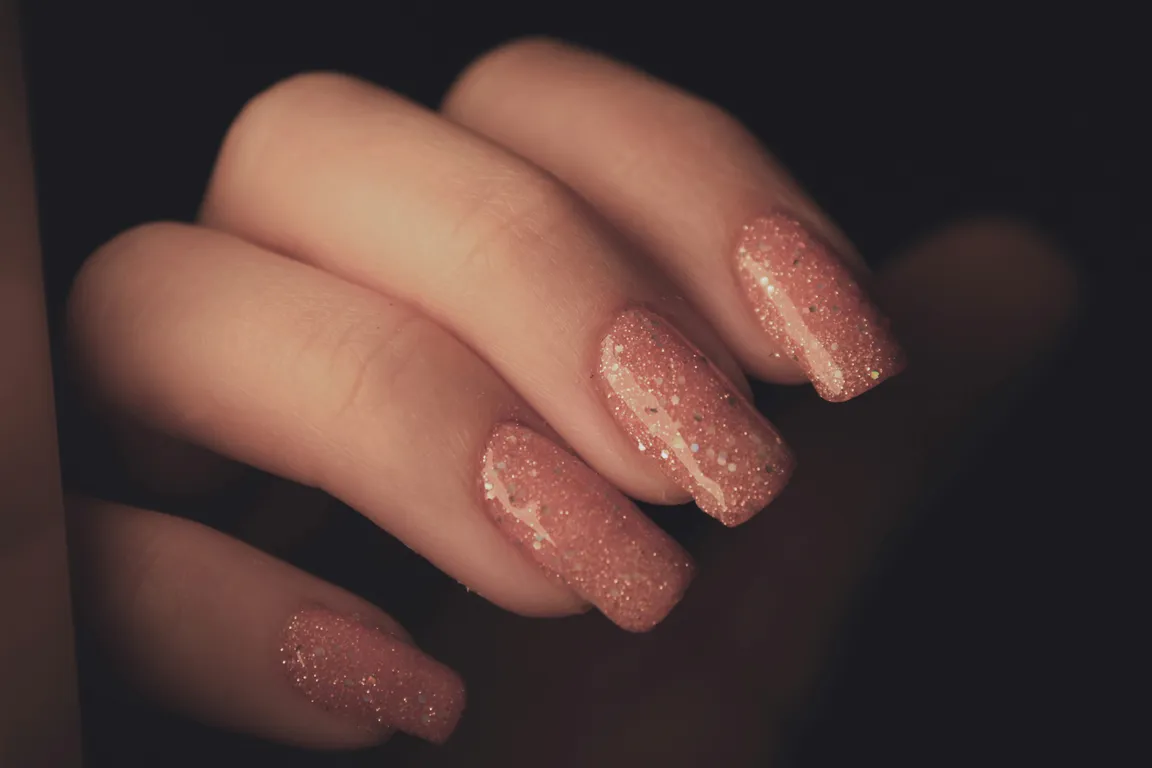 A hand with long square nails in rose pink packed with fine silver glitter, against a dark background