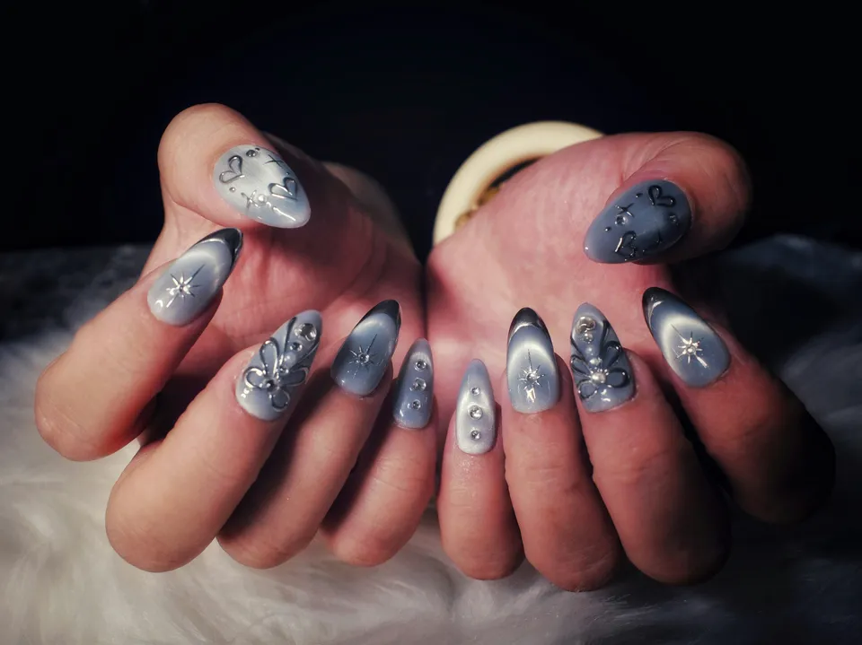 Two hands with grey-blue almond nails, sculpted silver flowers, hand-painted starbursts and crystal accents