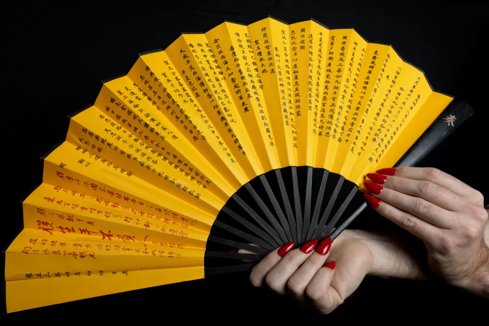 A hand with glossy red almond nails holding open a yellow silk fan covered in black and red calligraphy